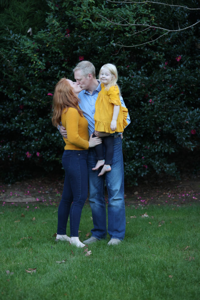Standing outside in front of Camelia bush wife kissing husband while he holds young daughter
what to wear for branding photos https://caryintegrativehealth.com 